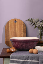 Purple textured bowl on a wooden surface with a cutting board and eggs against a purple wall.