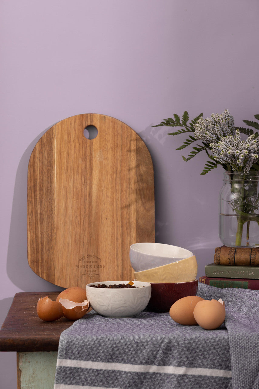 Wooden cutting board, bowls, and eggs on a table with a purple wall background