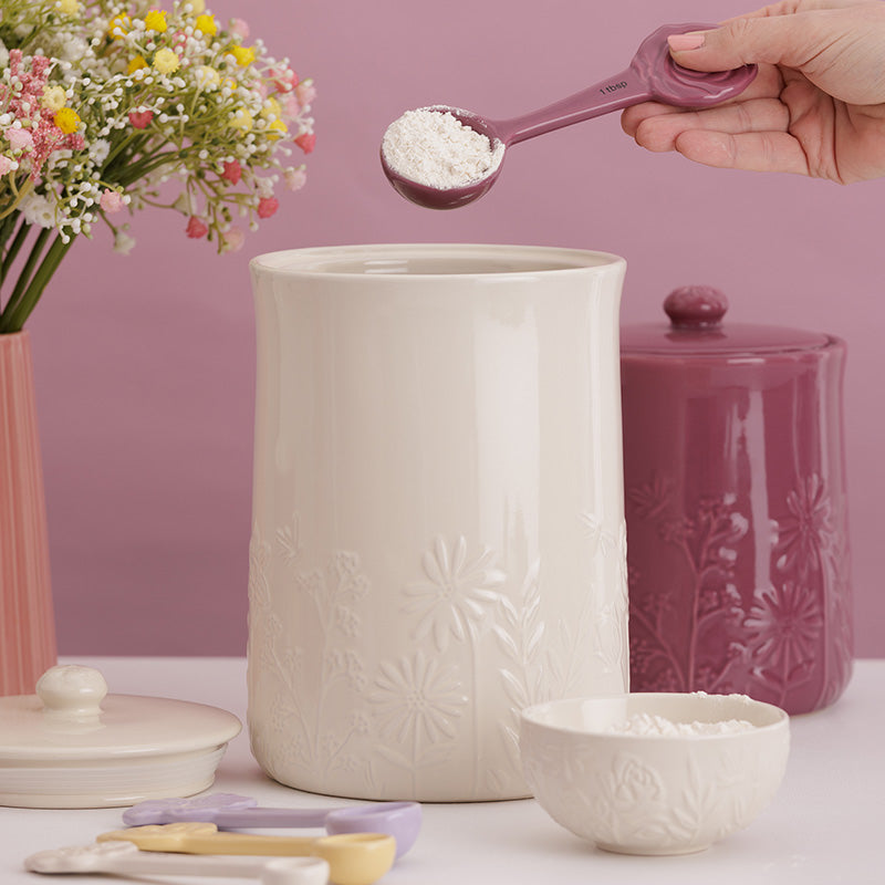 Person measuring flour with a spoon over a white container on a pink background